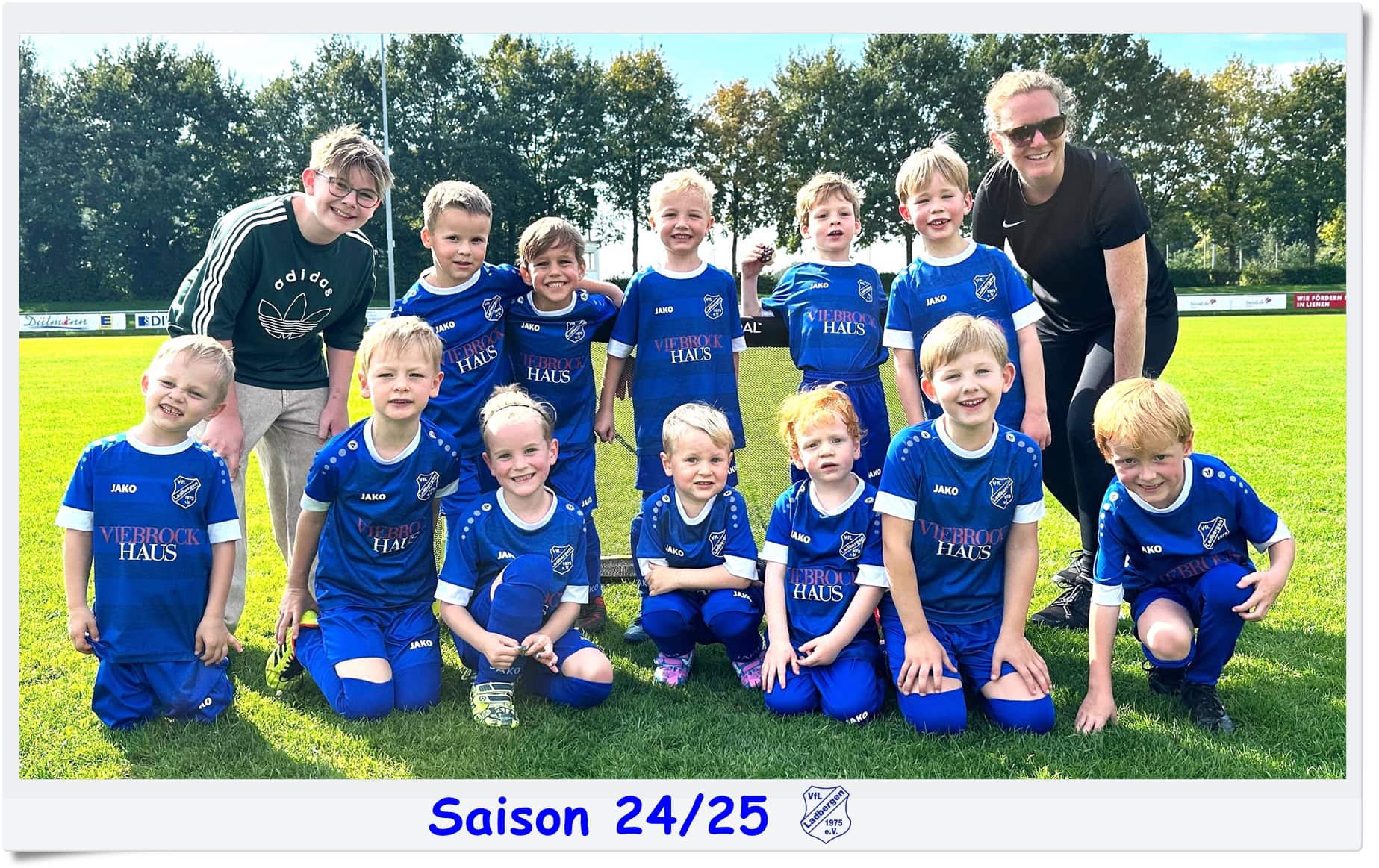 Kinderfußballmannschaft in blauen Trikots, draußen lächelnd, mit zwei Erwachsenen; Bäume im Hintergrund.